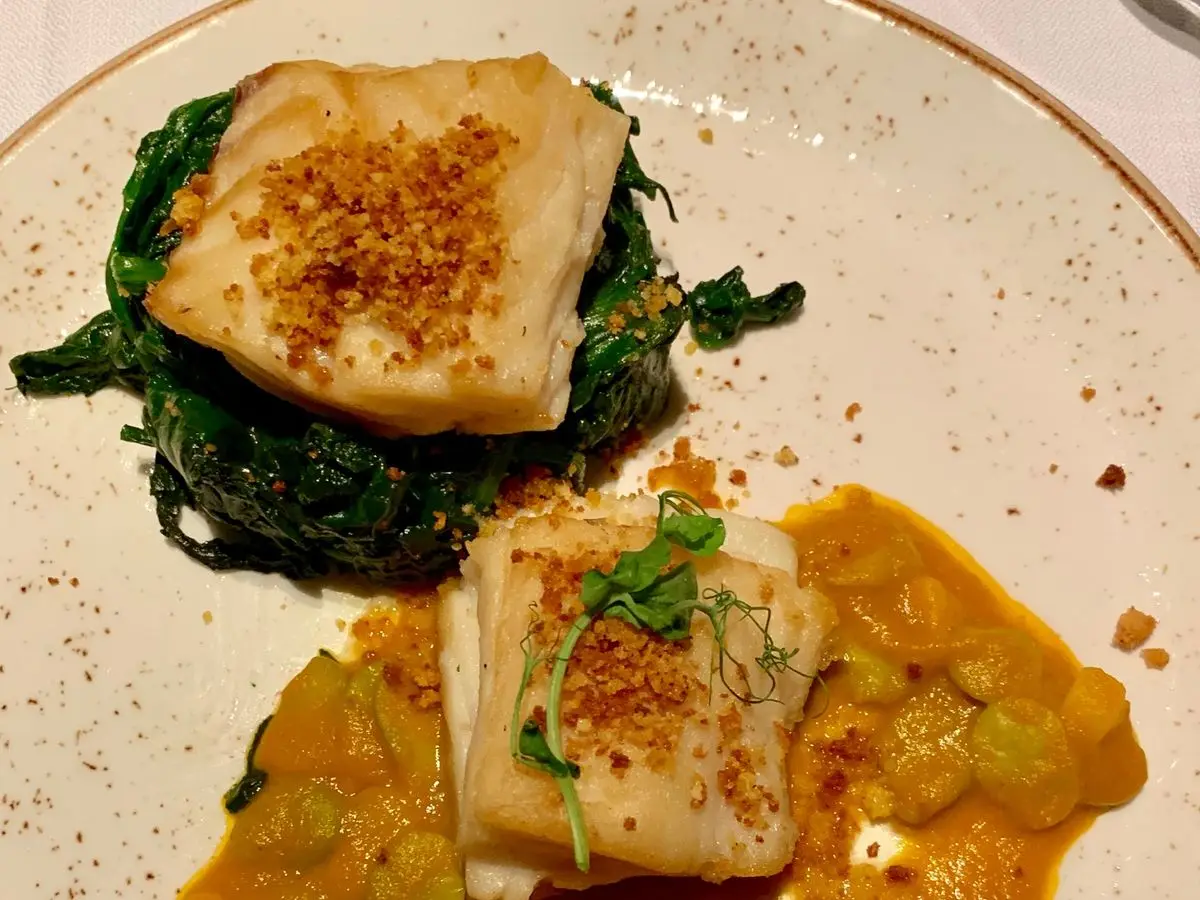 Dos lomos de pescado blanco con migas tostadas sobre espinacas y habas en salsa anaranjada, en un plato claro