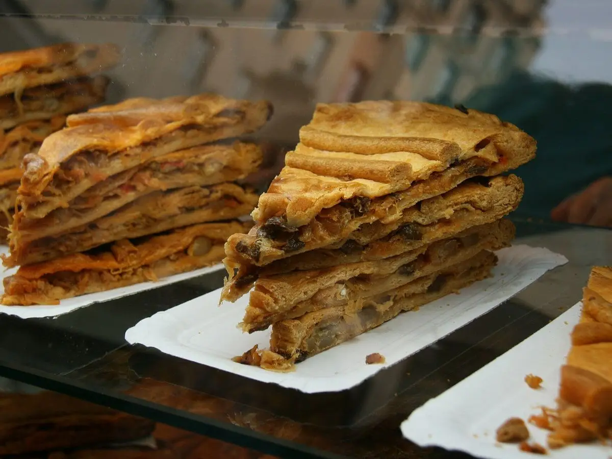 Porciones de empanada gallega de masa dorada y hojaldrada sobre bandejas de cartón blancas