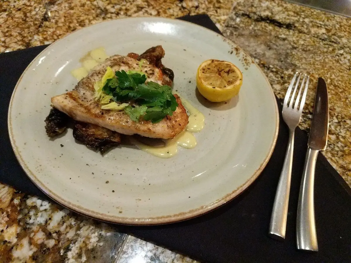 Filete de pez espada a la plancha con hierbas y medio limón en un plato blanco con cubiertos