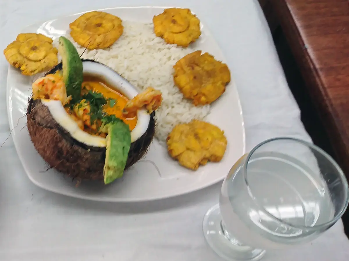 Encocado servido en un coco con arroz blanco, patacones fritos y aguacate en un plato blanco
