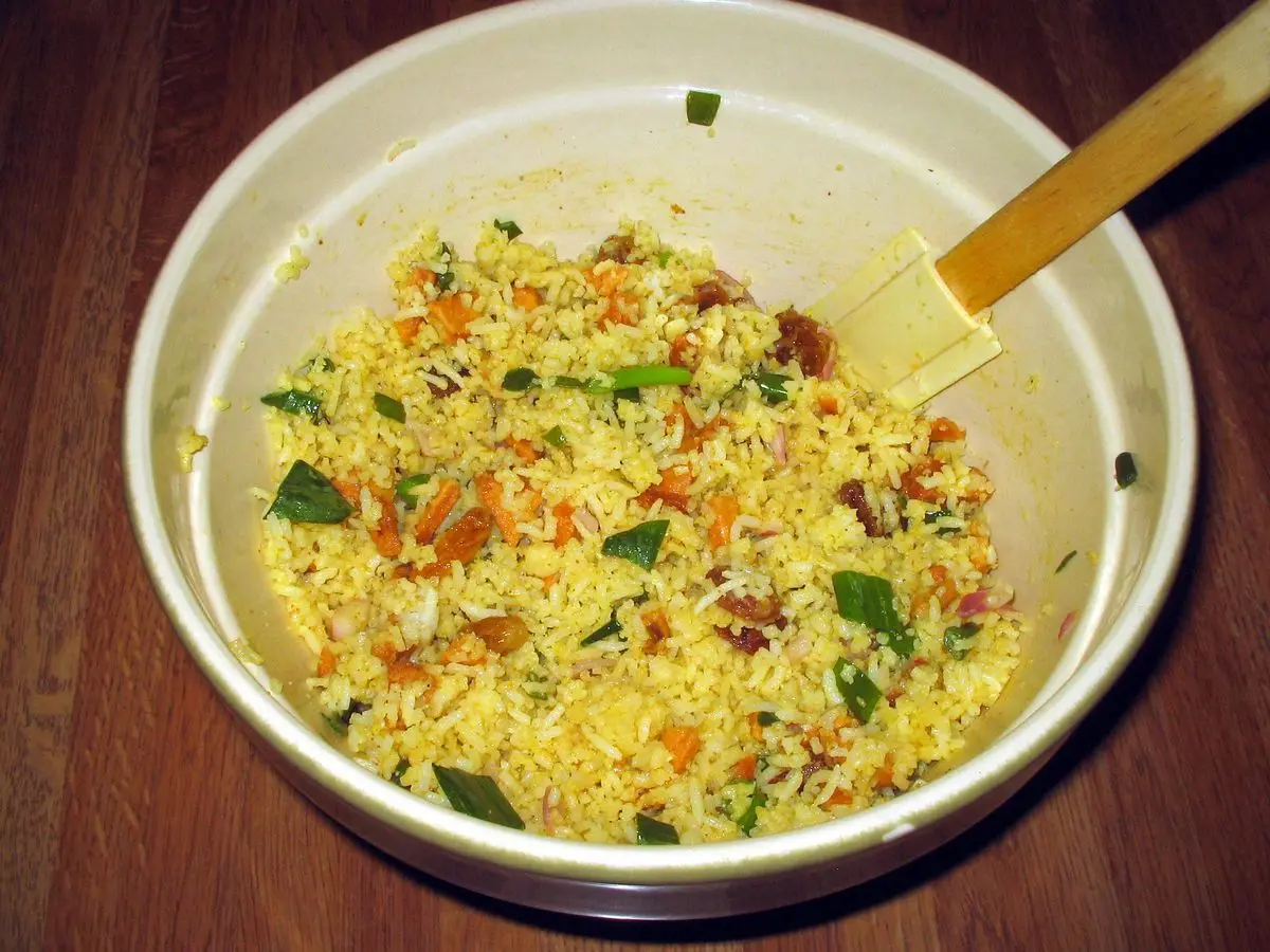 Cuenco claro de ensalada de arroz amarillo al curry con trocitos de fruta y hierbas, con una espátula