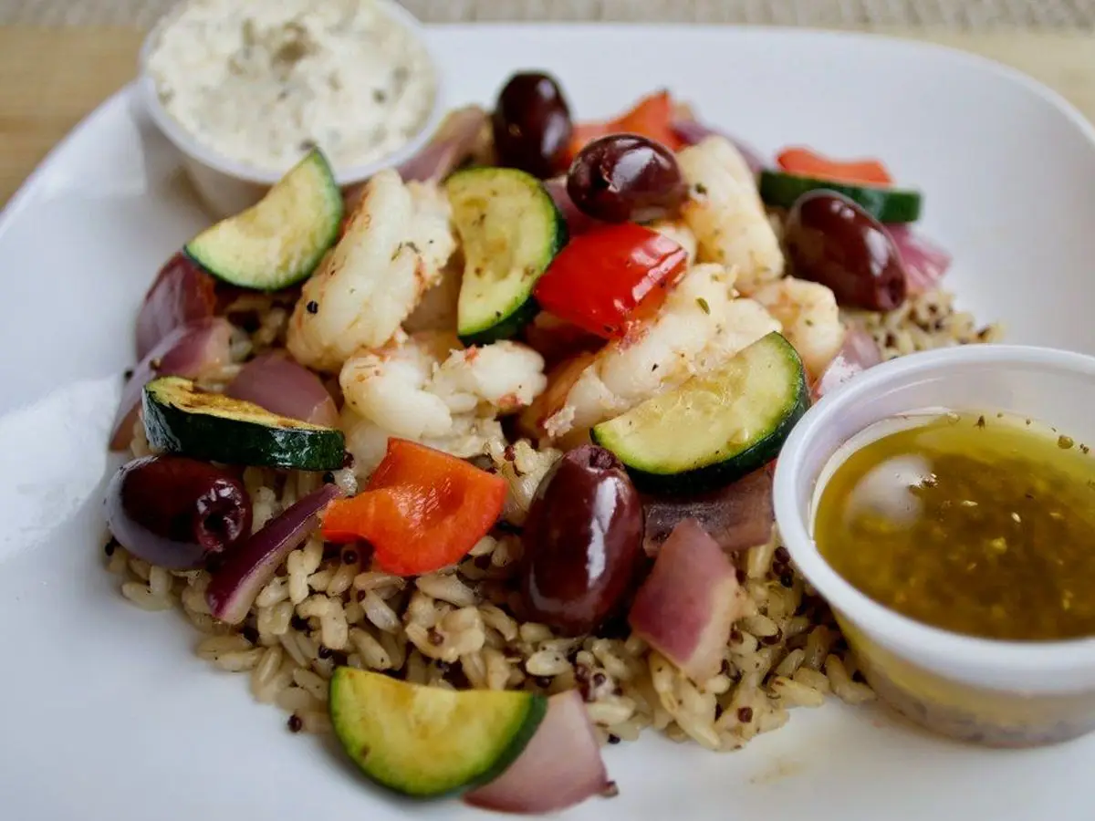Plato de ensalada de arroz integral con langostinos, calabacín en rodajas, pimiento y aceitunas, con salsa al lado