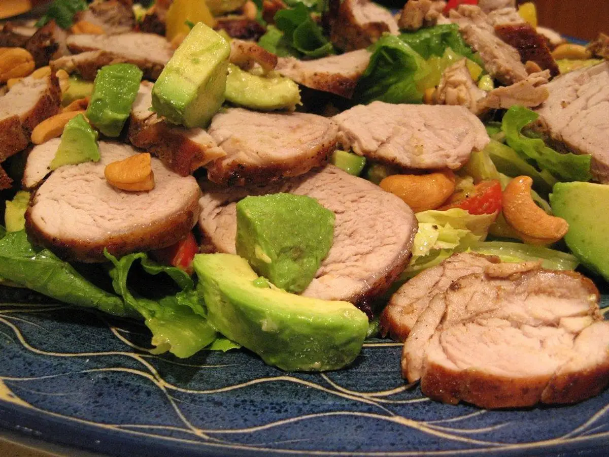 Ensalada de hojas verdes con rodajas de solomillo de cerdo, aguacate y frutos secos en un plato azul