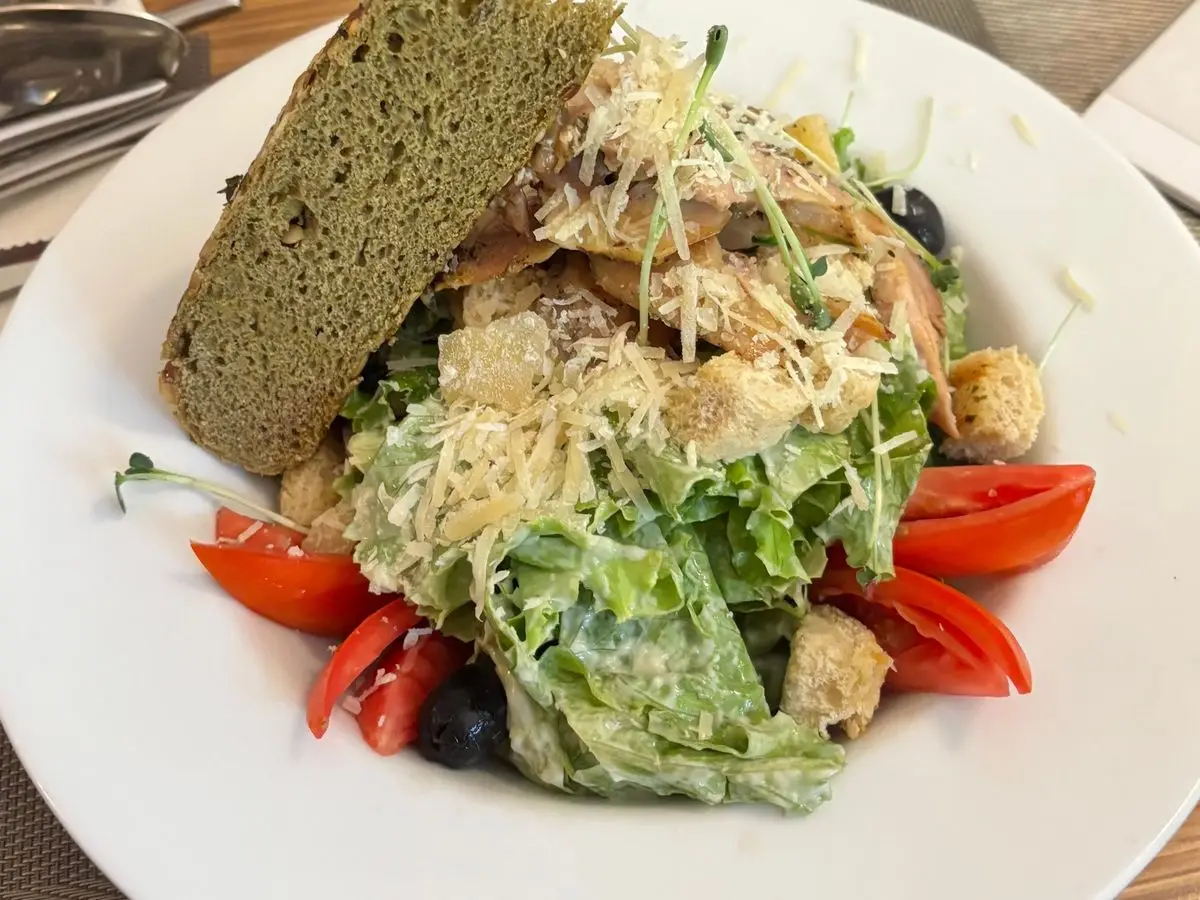 Ensalada verde con pollo, picatostes, queso rallado, aceitunas y tomate, junto a una rebanada de pan
