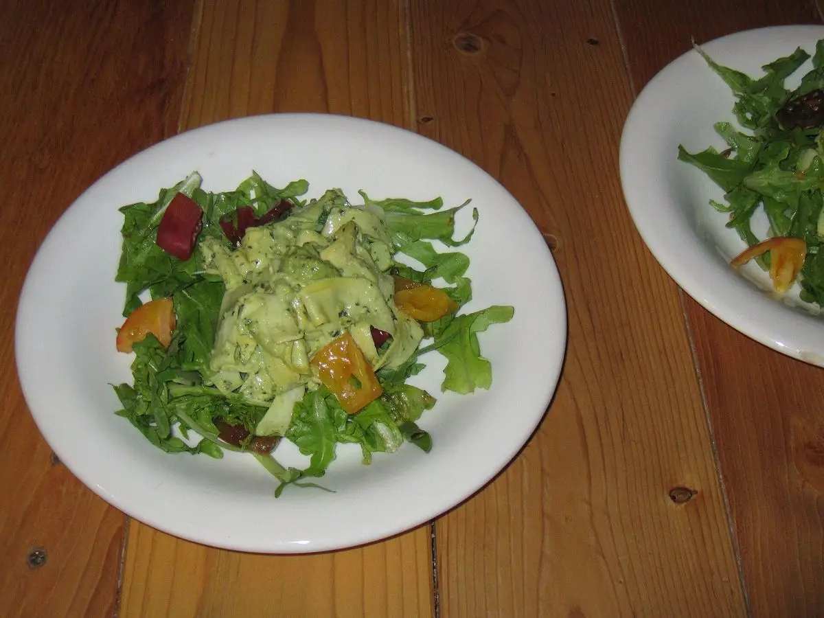 Plato blanco con ensalada de hojas verdes, aguacate aliñado y trozos de naranja sobre mesa de madera
