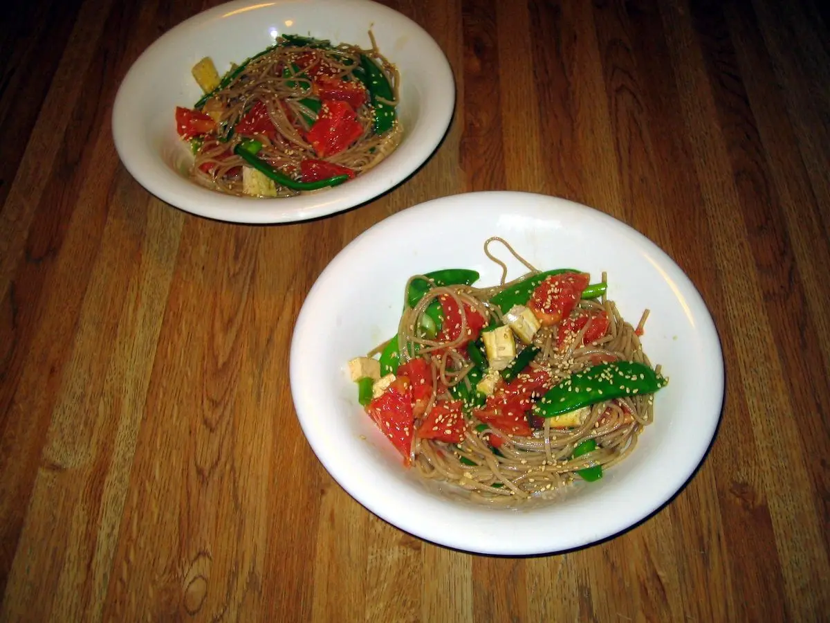 Dos platos blancos con fideos de soba salteados con verduras rojas y verdes sobre una mesa de madera
