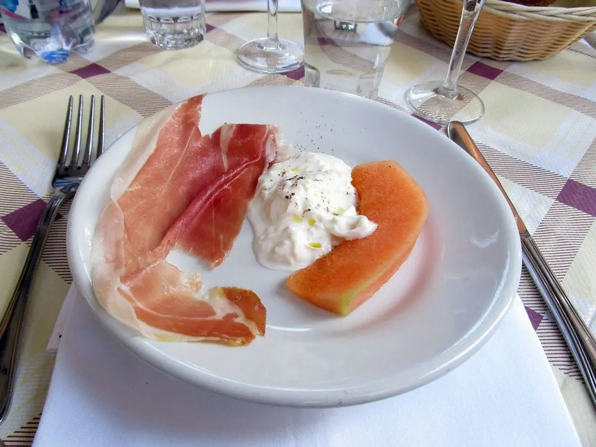 Plato blanco con jamón curado, melón en gajos y queso blanco cremoso sobre un mantel