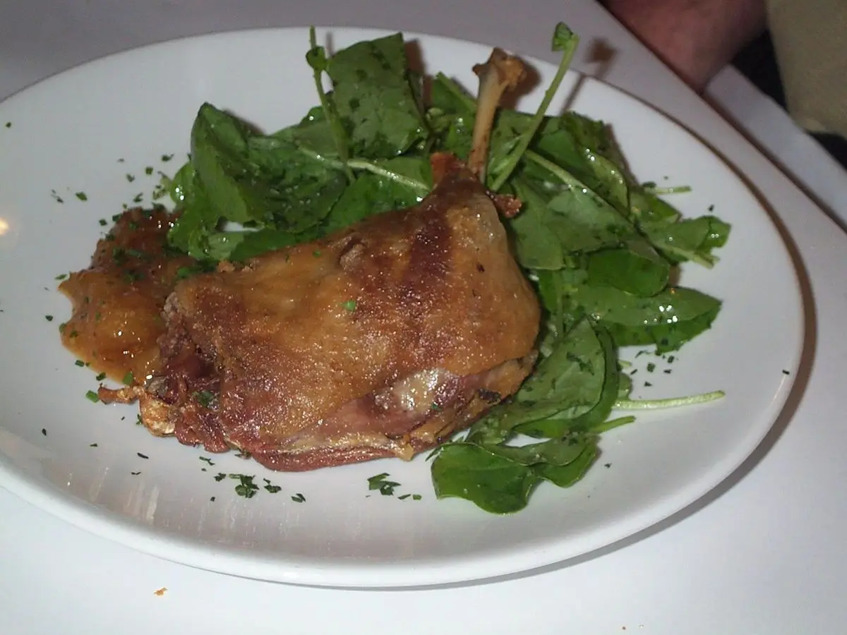 Muslo de pato confitado dorado sobre hojas verdes de ensalada en un plato blanco