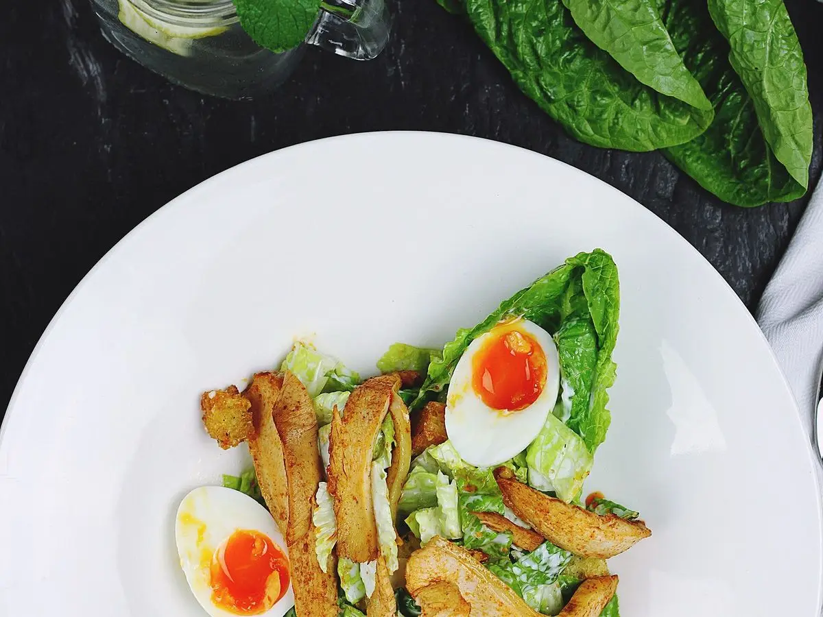 Plato blanco con ensalada de hojas verdes, pollo, medio huevo cocido y picatostes