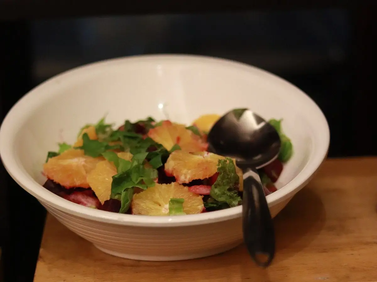 Cuenco blanco de ensalada con gajos de naranja, hojas verdes y remolacha asada sobre una mesa de madera