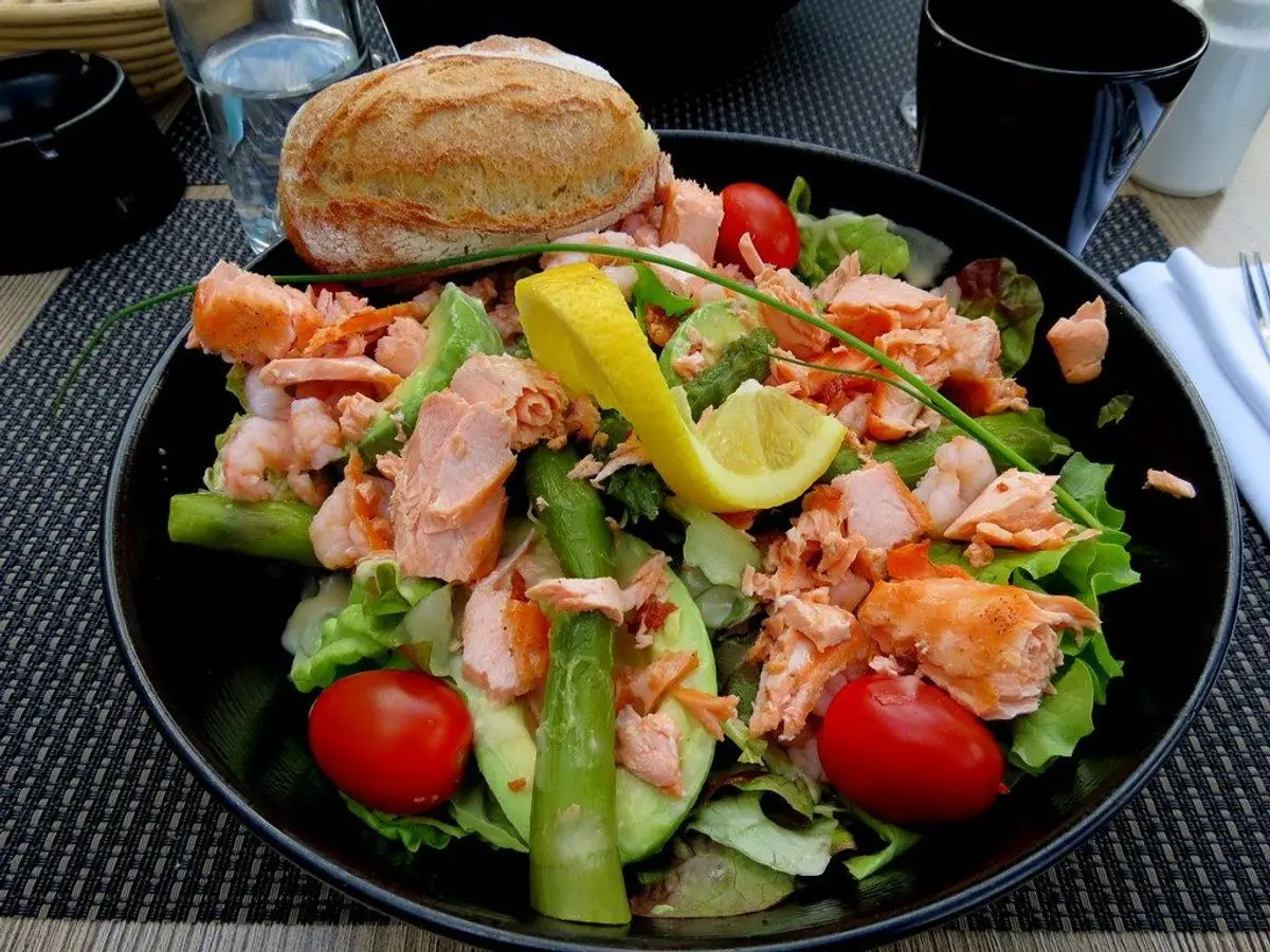 Ensalada en plato negro con salmón desmenuzado, tomates cherry, hojas verdes, espárragos y rodajas de limón