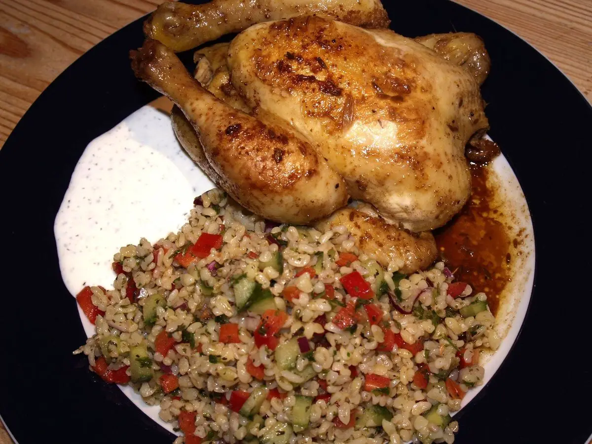 Pollo asado dorado junto a una ensalada de trigo con tomate, pepino y hierbas sobre un plato oscuro