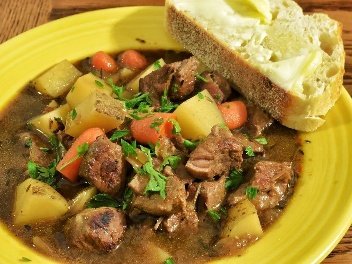 Plato hondo amarillo con guiso de cordero, patata, zanahoria y perejil junto a una rebanada de pan