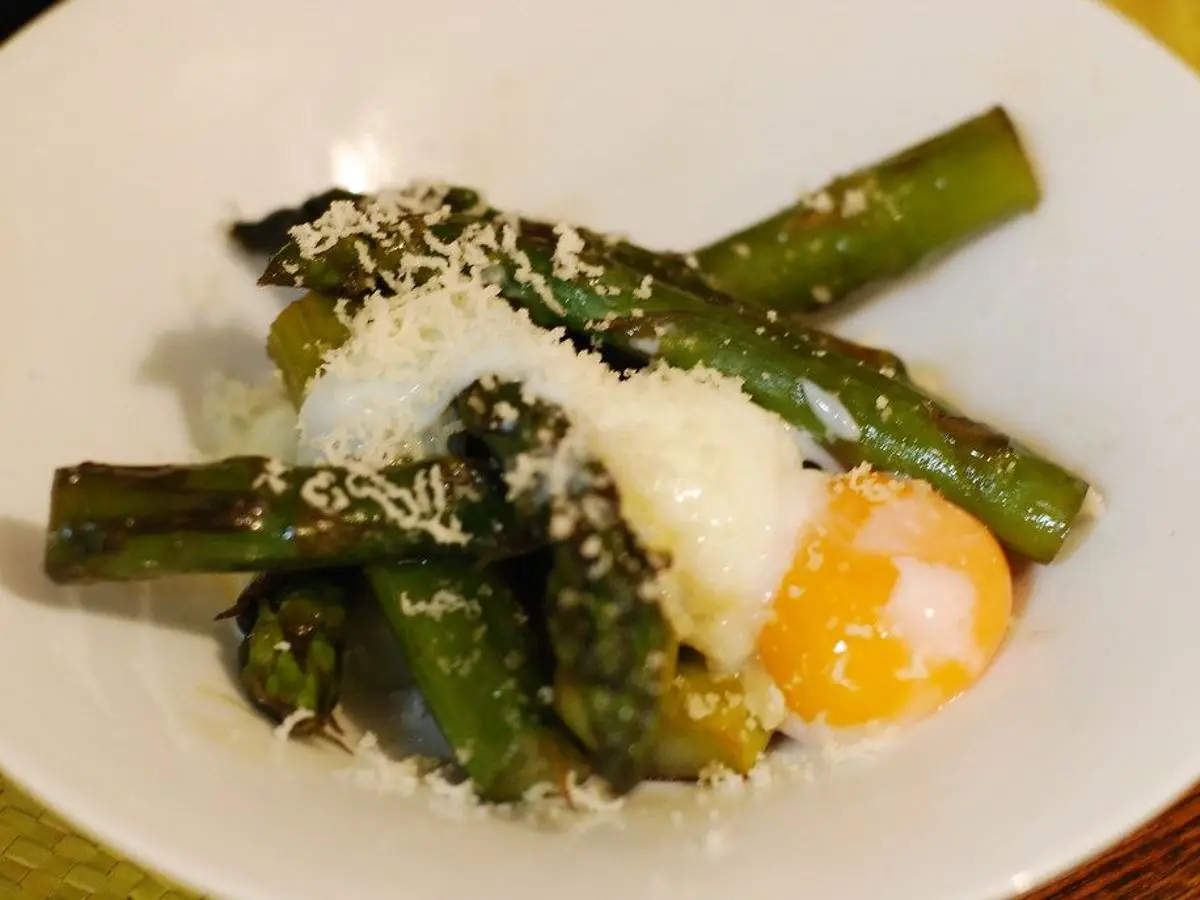 Espárragos verdes a la plancha con un huevo de yema líquida y queso rallado en un plato blanco