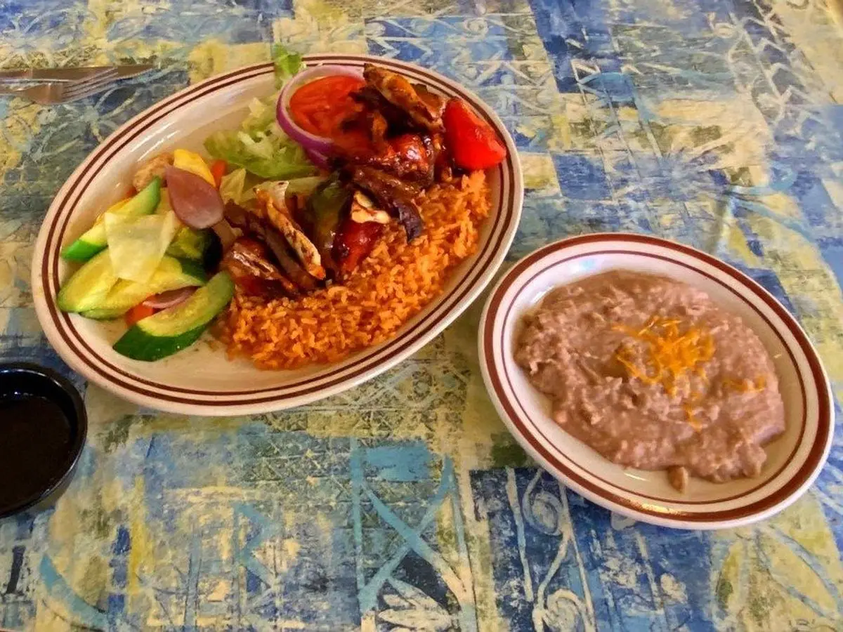 Plato con carne a la parrilla, arroz rojo, ensalada de pepino y aguacate junto a un plato de frijoles