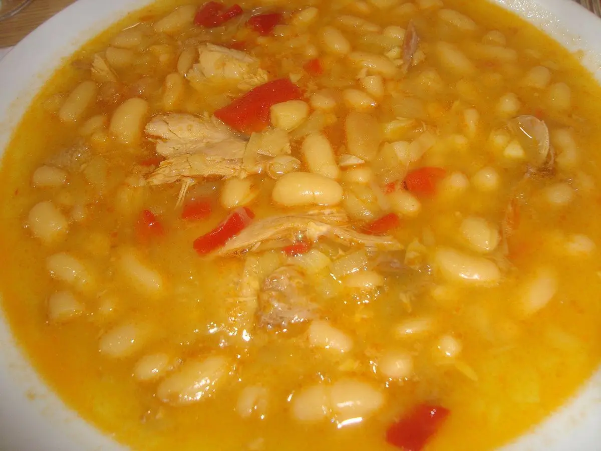 Plato de alubias blancas guisadas con carne de caza desmigada y tiras de pimiento rojo en caldo anaranjado