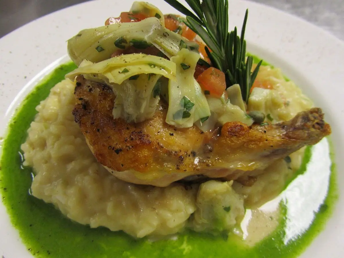 Risotto cremoso con alcachofa laminada, pollo dorado, ramita de romero y salsa verde en un plato blanco