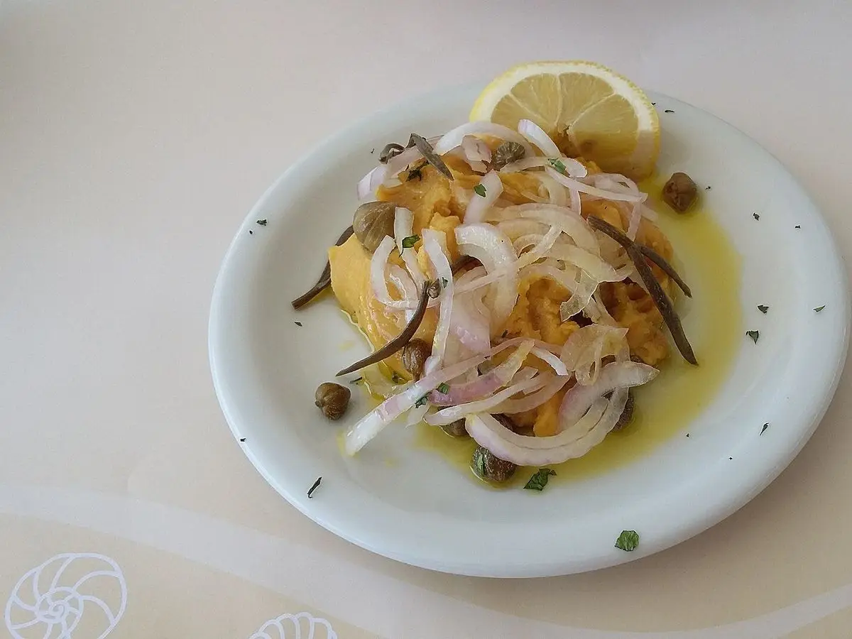 Plato blanco con puré de legumbre amarillo cubierto de aros de cebolla, alcaparras y una rodaja de limón