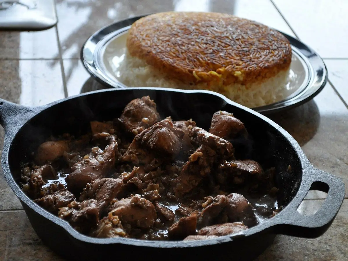 Guiso persa oscuro de carne en sartén de hierro junto a un plato de arroz con costra dorada