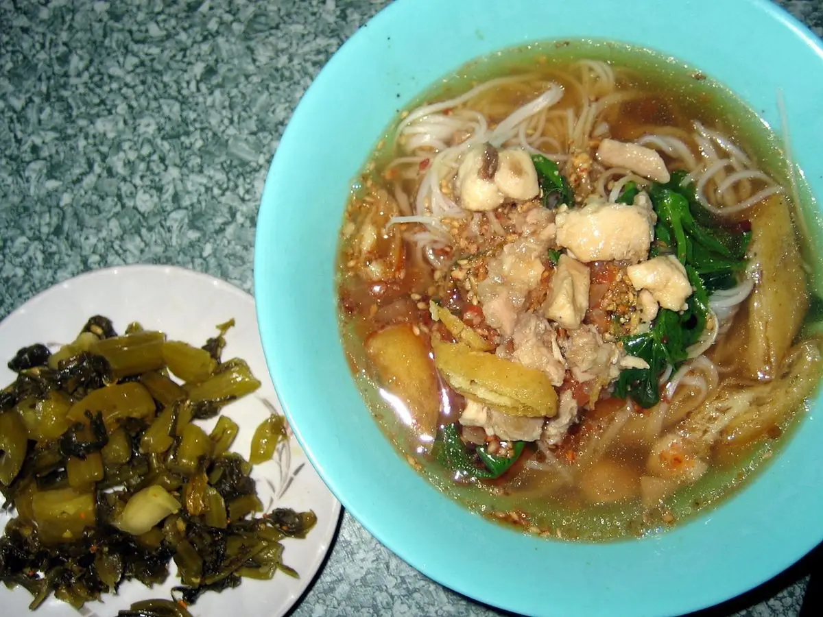 Cuenco turquesa de fideos de arroz en caldo con pollo y verduras, con encurtido verde en un plato aparte