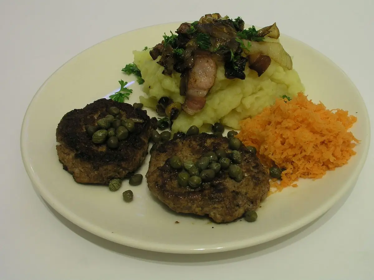 Dos hamburguesas de carne con alcaparras sobre puré de patata y zanahoria rallada