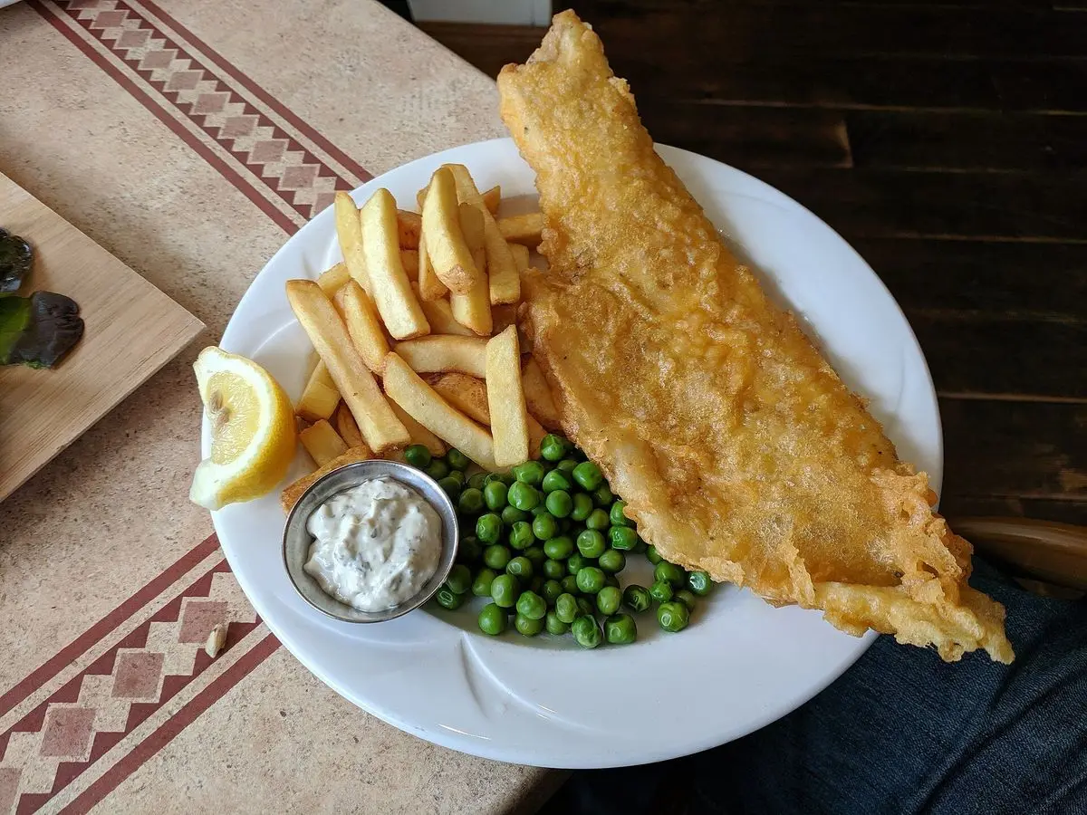 Filete de pescado rebozado con patatas fritas, guisantes y salsa blanca en un plato
