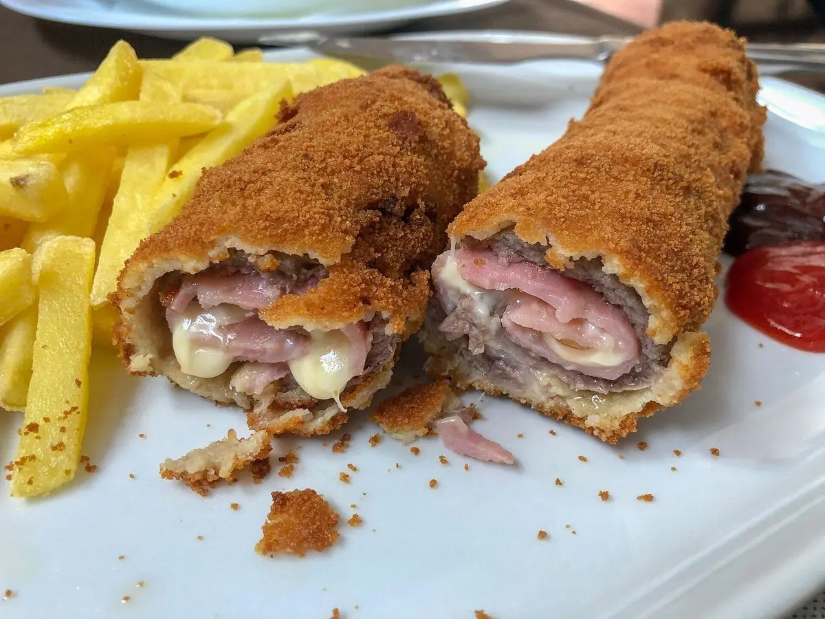 Flamenquín empanado cortado por la mitad que muestra el jamón y el queso, con patatas fritas