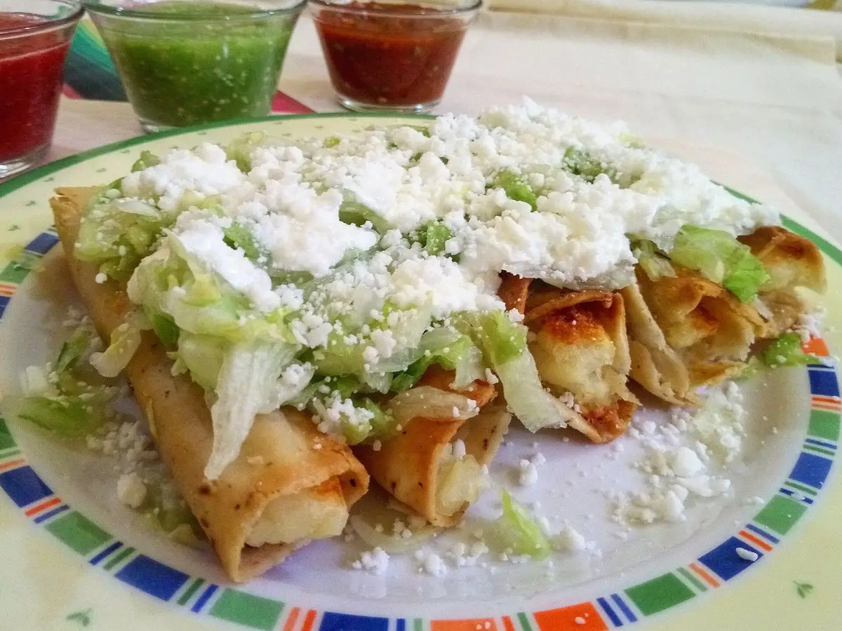Flautas doradas cubiertas de lechuga rallada y queso, con pequeños cuencos de salsa al fondo