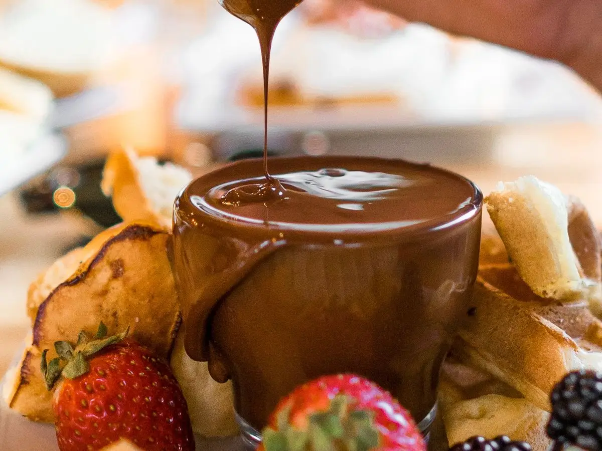 Fondue de chocolate negro cayendo sobre fresas, moras y trozos de fruta y bizcocho