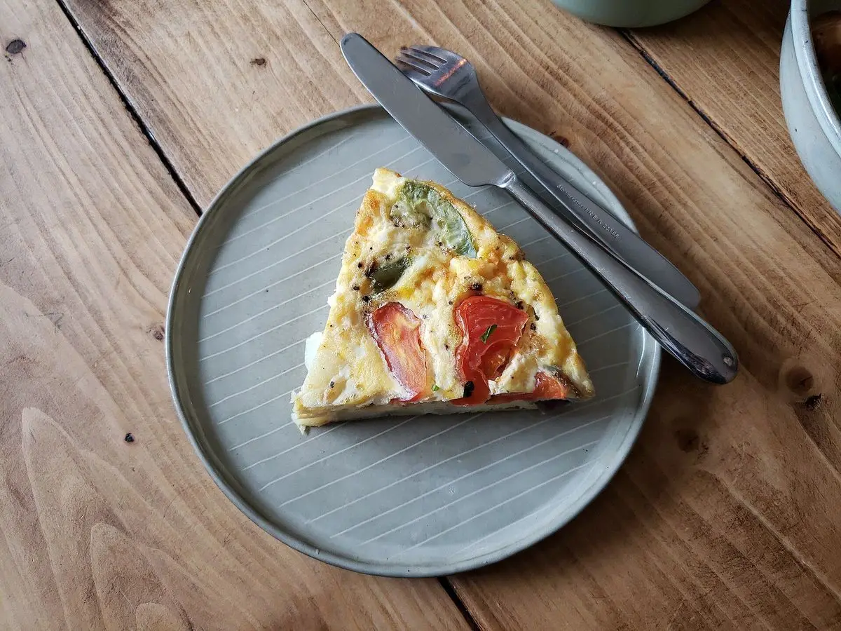 Porción de frittata con pimiento y verduras en un plato gris sobre una mesa de madera, con cuchillo y tenedor