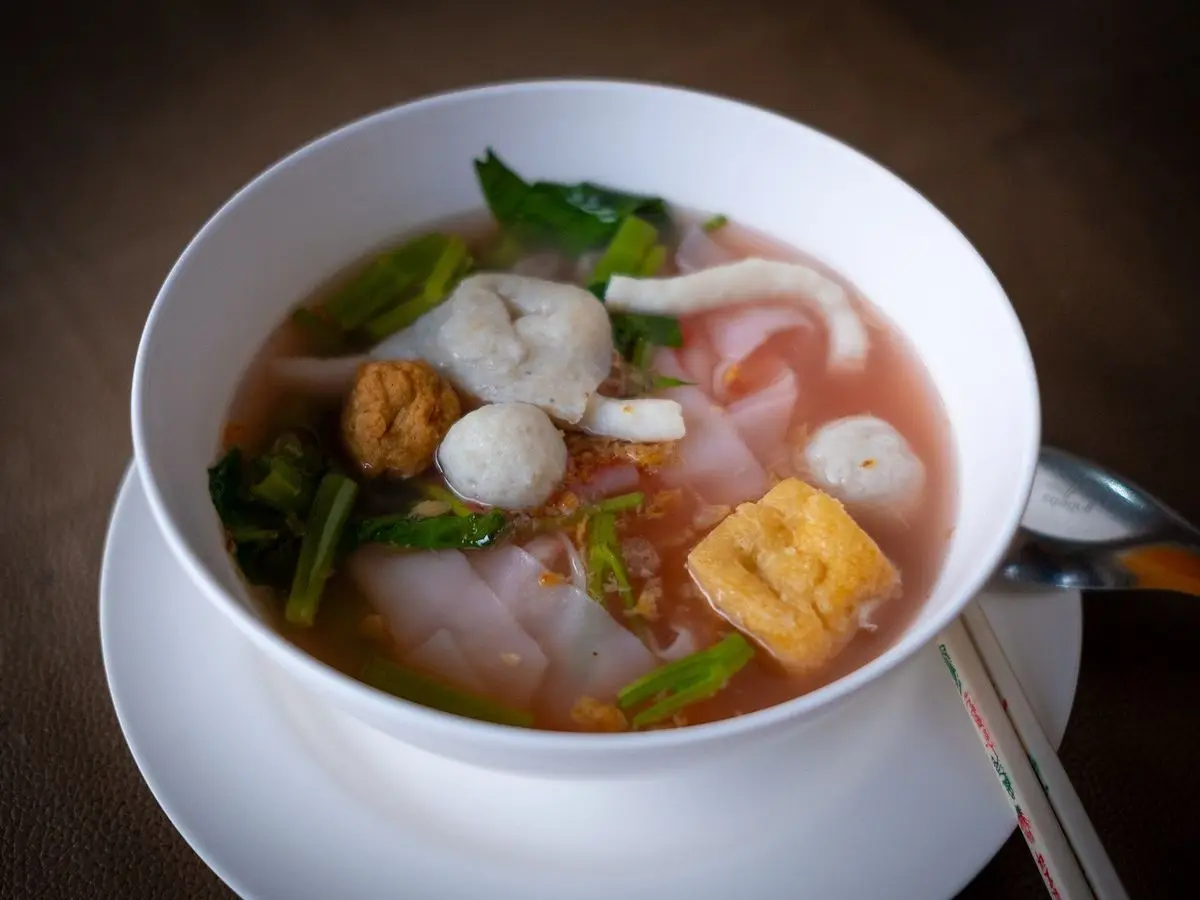 Cuenco blanco de sopa clara con bolas de pescado, tofu frito, fideos y verdura verde