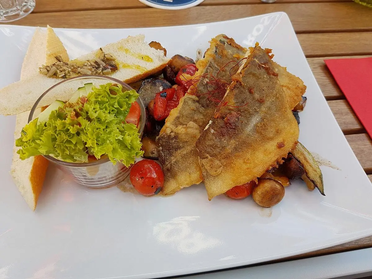 Filete de pescado dorado con tomates cherry y verduras salteadas en un plato cuadrado blanco, con ensalada