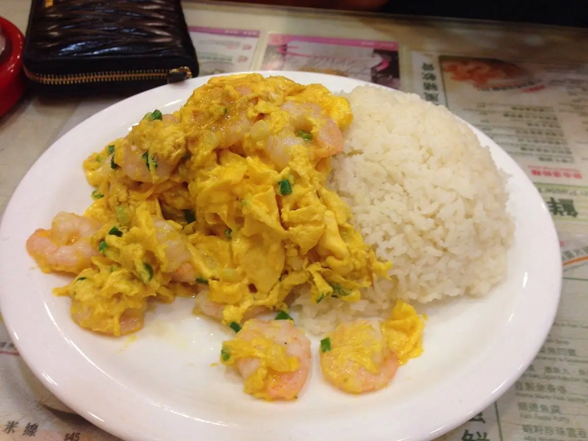Plato blanco de huevo revuelto con gambas y una porción de arroz blanco sobre un mantel de papel