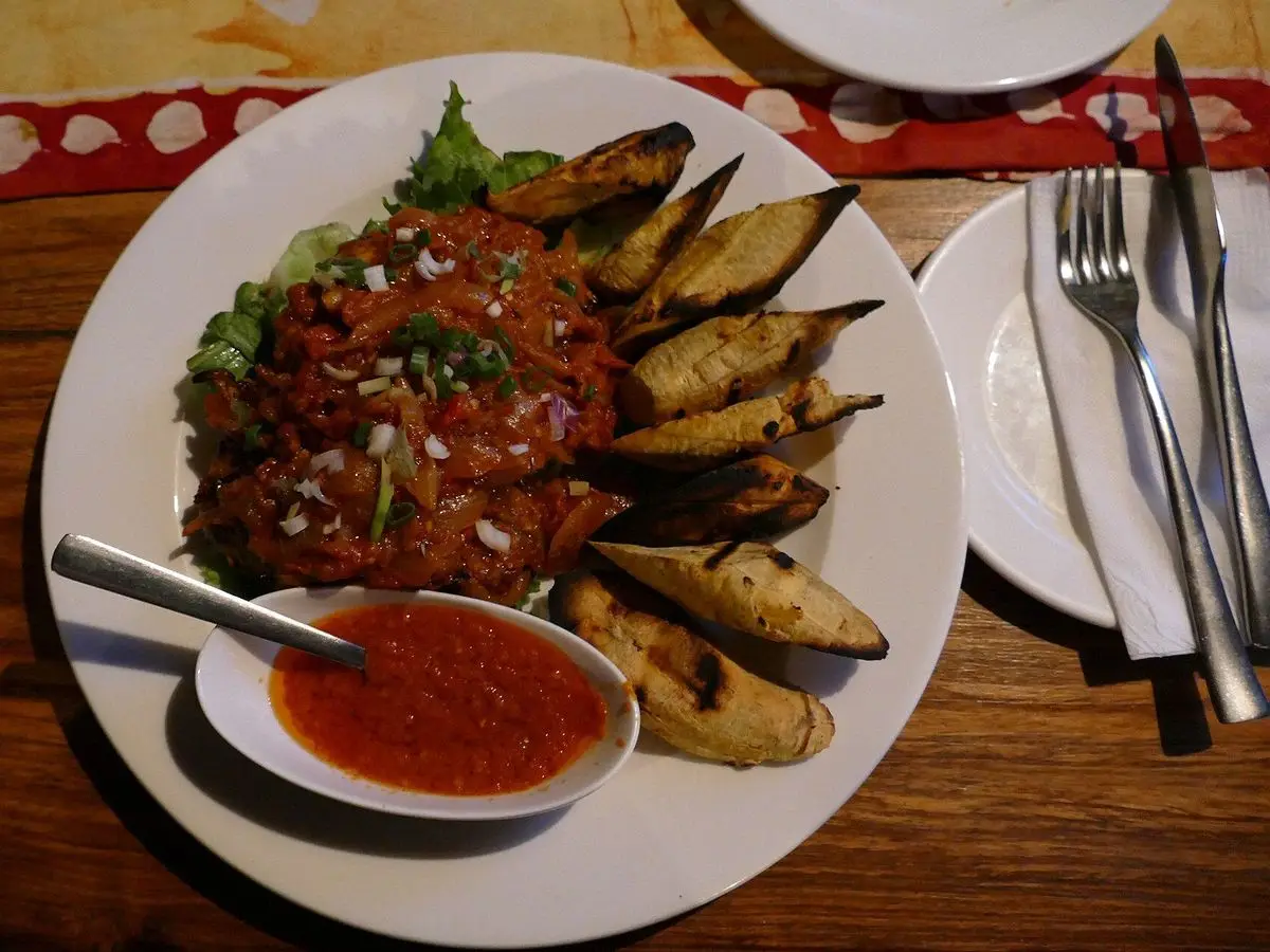 Plato con pollo en salsa de pimientos rojos y plátano macho asado en rodajas, con un cuenco de salsa
