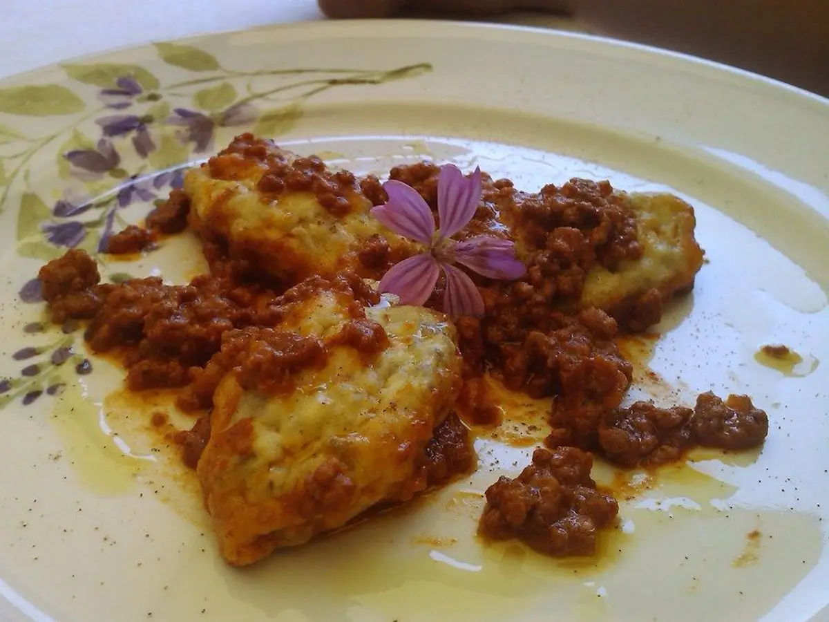 Gnudi dorados en un plato con flores, cubiertos de salsa rojiza y una flor morada de adorno