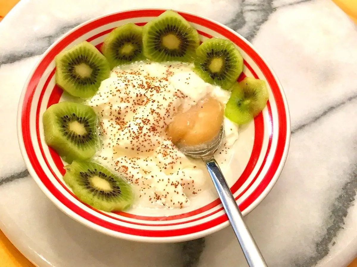 Cuenco de rayas rojas con yogur espeso, rodajas de kiwi alrededor, semillas espolvoreadas y una cuchara