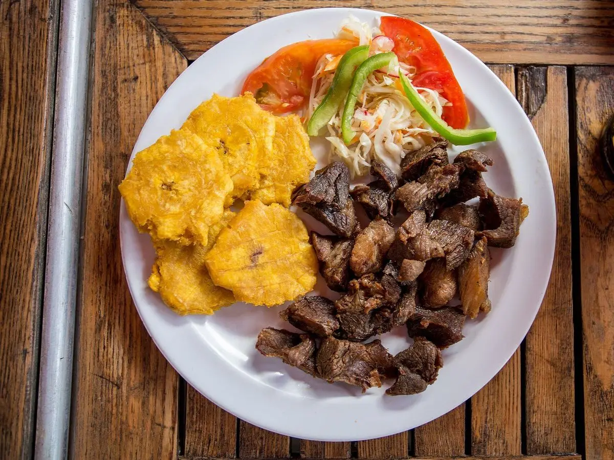 Plato con cerdo frito en dados, plátano verde frito y ensalada de pikliz sobre mesa de madera