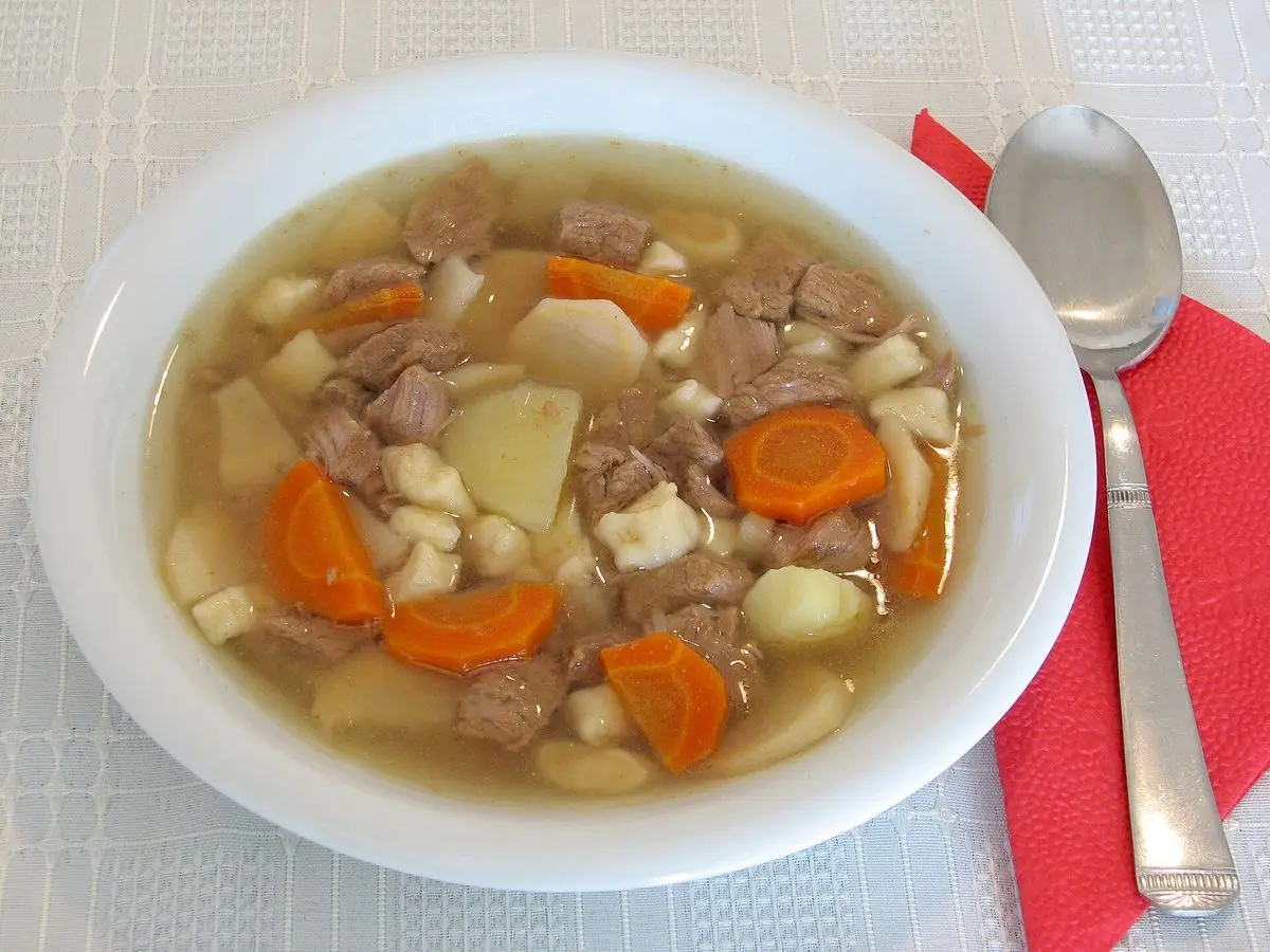 Plato hondo blanco de sopa gulash con patata, zanahoria y legumbres sobre un mantel claro con cuchara