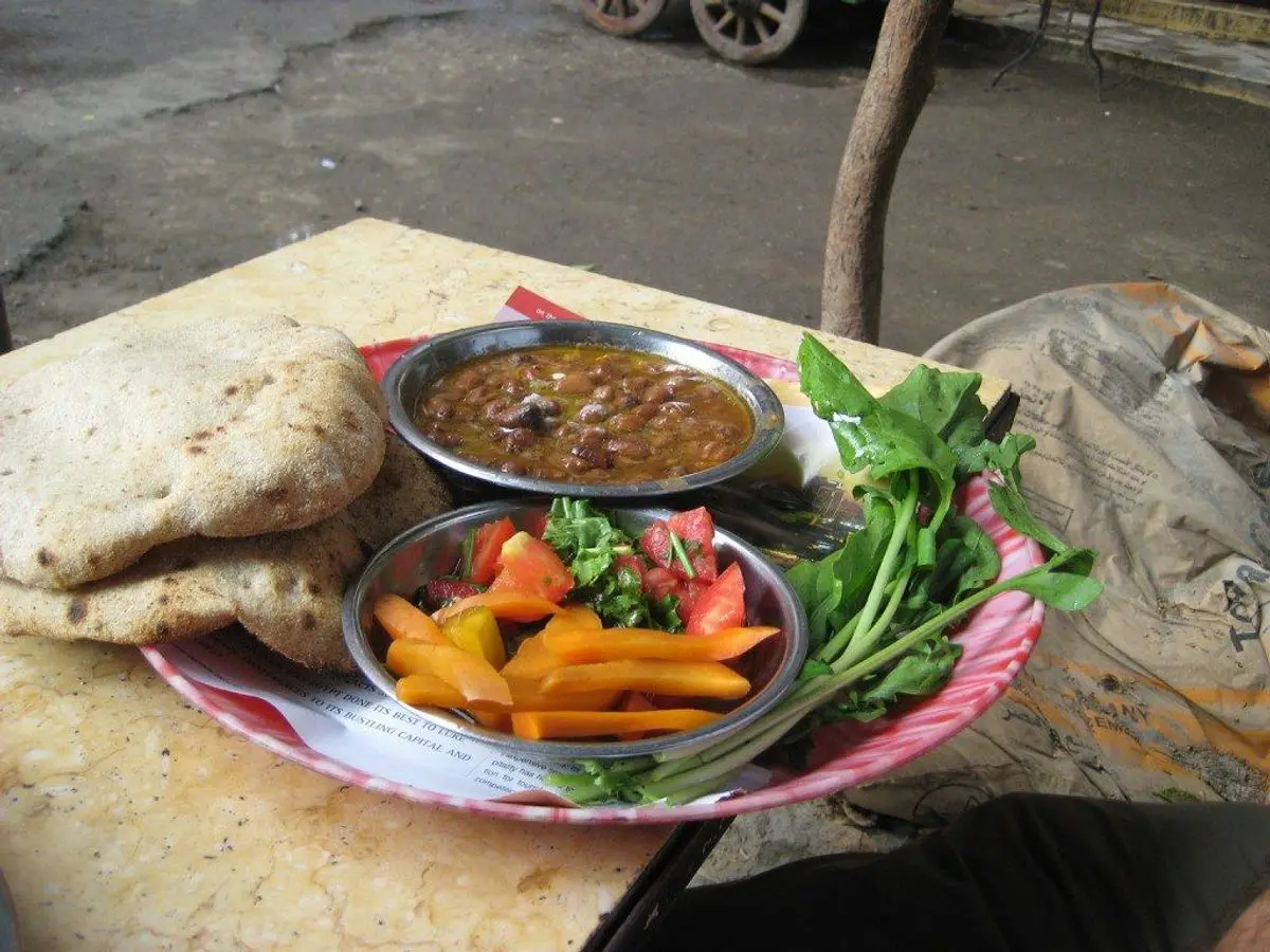 Plato con cuenco de ful medames, pan de pita, rodajas de zanahoria, tomate y hojas verdes sobre una mesa