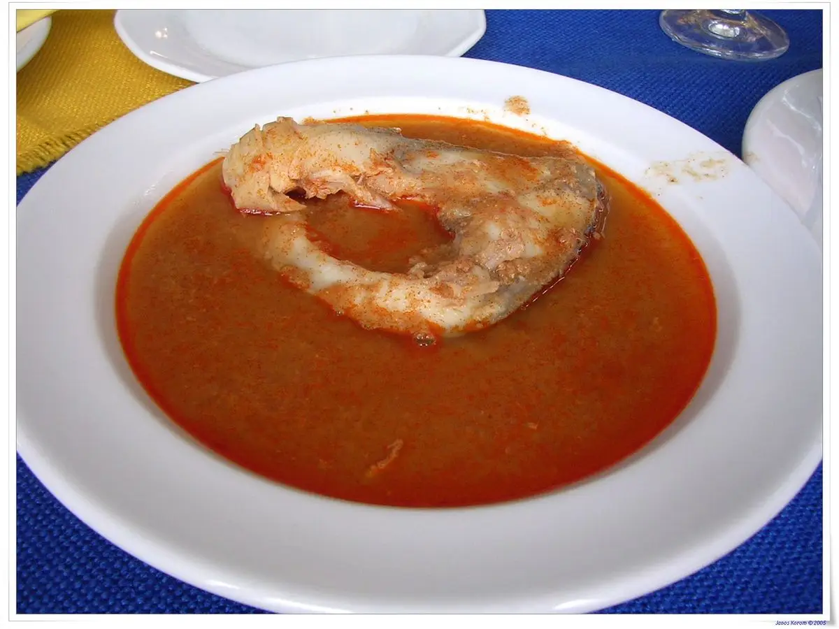 Plato hondo de sopa de pescado rojiza al pimentón con un trozo de pescado, sobre un mantel azul