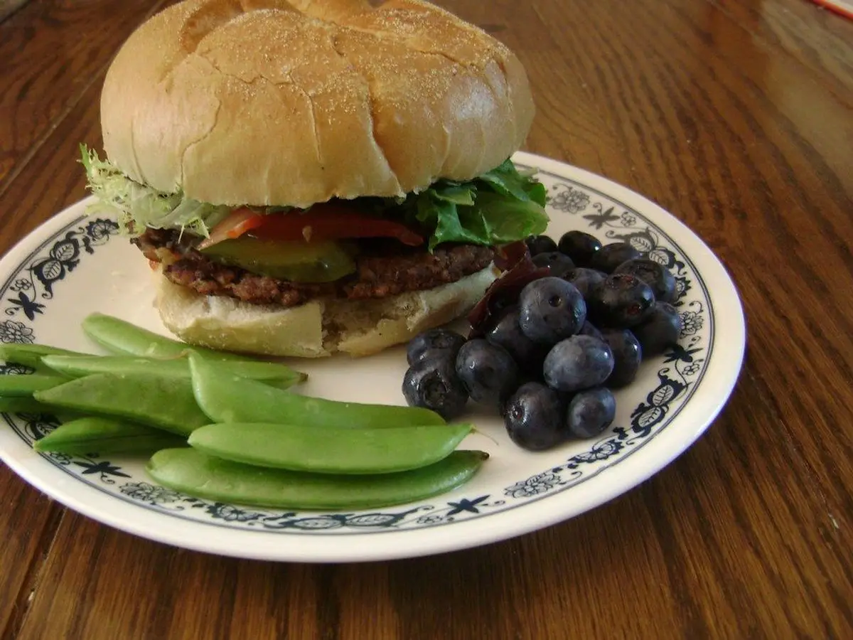 Hamburguesa de alubias negras en pan con lechuga, junto a guisantes tirabeques y arándanos en un plato
