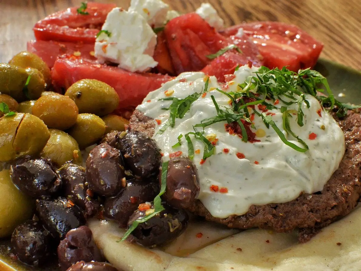 Hamburguesa de cordero sobre pan con salsa de yogur, tomate, aceitunas y hierbas