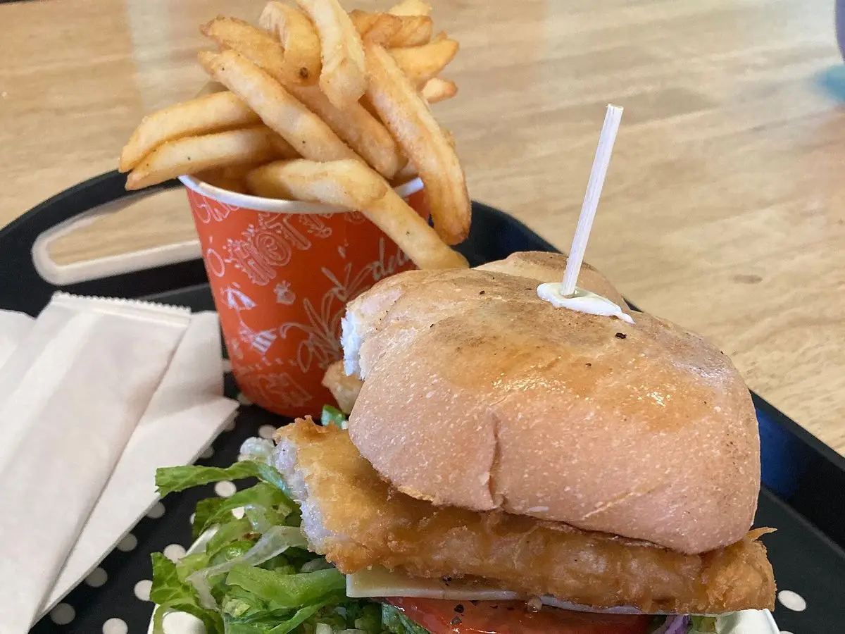 Hamburguesa de pescado en pan con lechuga, tomate y cebolla morada, servida sobre un plato con patatas