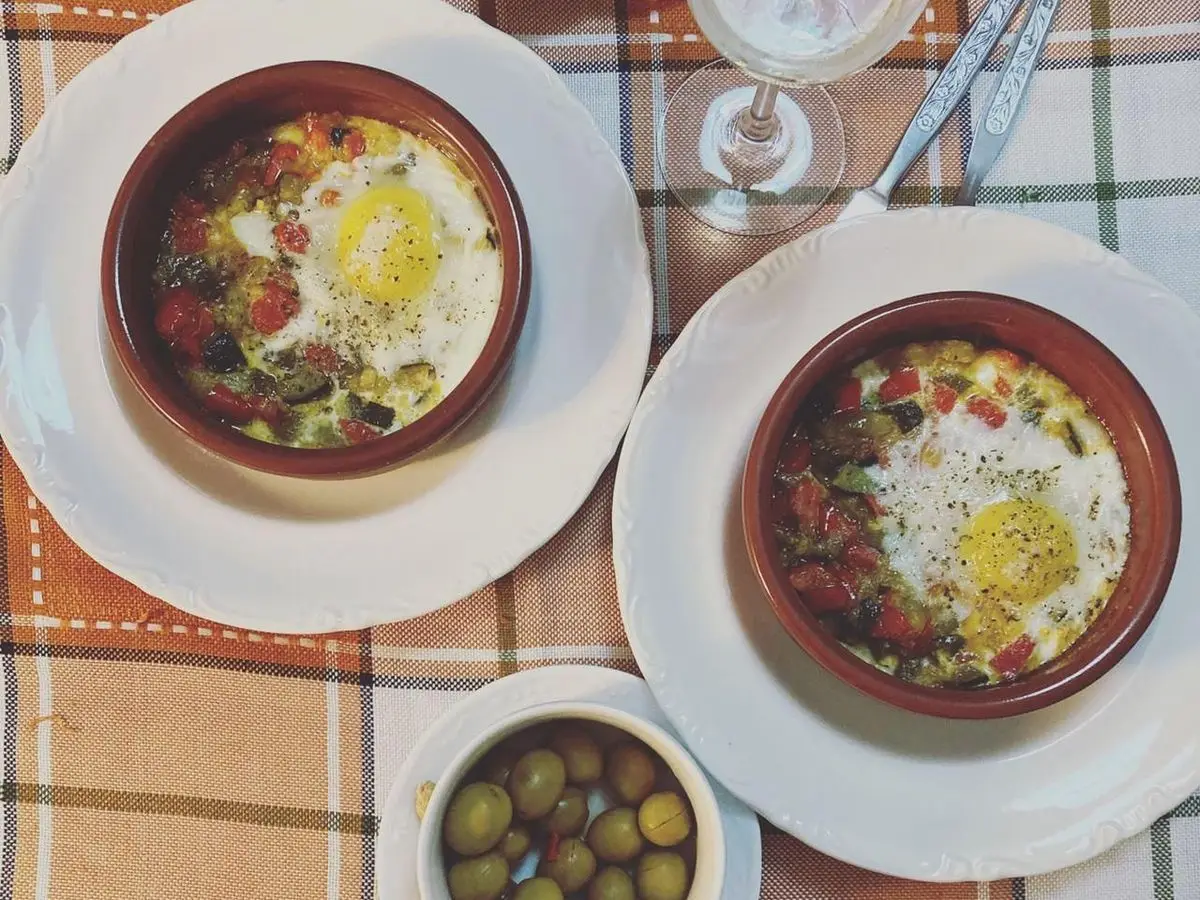Dos cazuelitas de barro con huevos cuajados al horno sobre sofrito de verduras en la mesa
