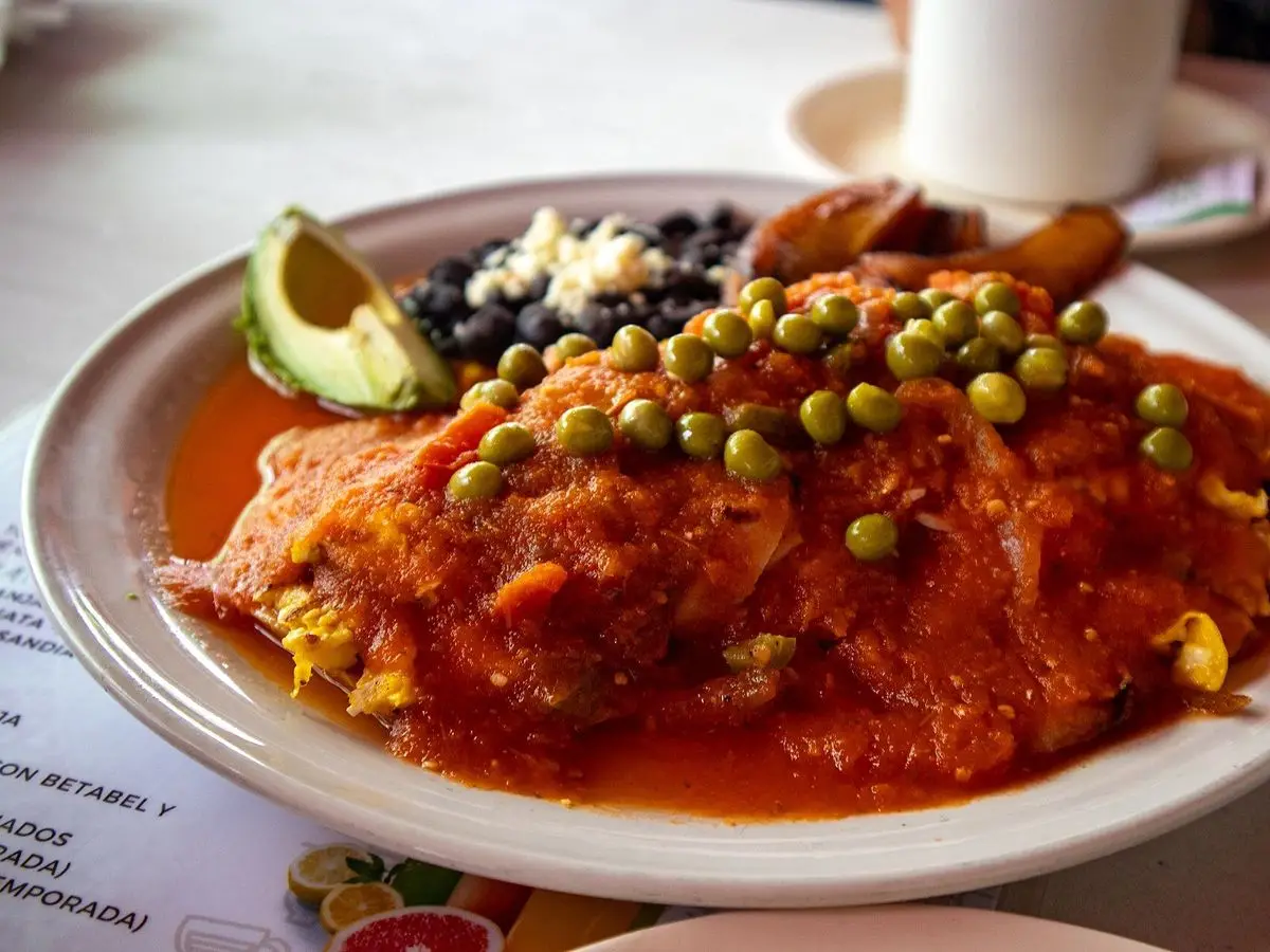 Huevos motuleños con salsa de tomate, guisantes, frijoles negros, aguacate y plátano frito