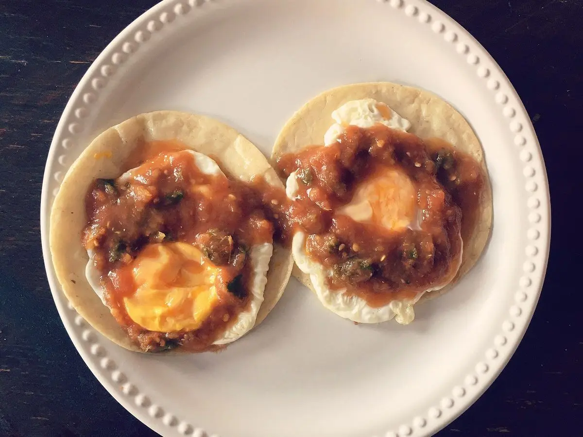 Dos tortillas de maíz con huevo frito, alubias y salsa roja sobre un plato blanco