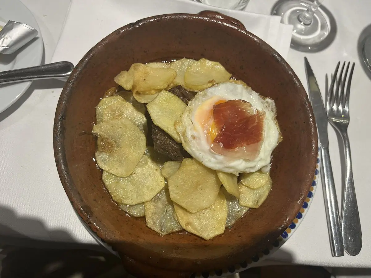 Cazuela de barro con patatas fritas en rodajas y un huevo frito coronado con una loncha de jamón