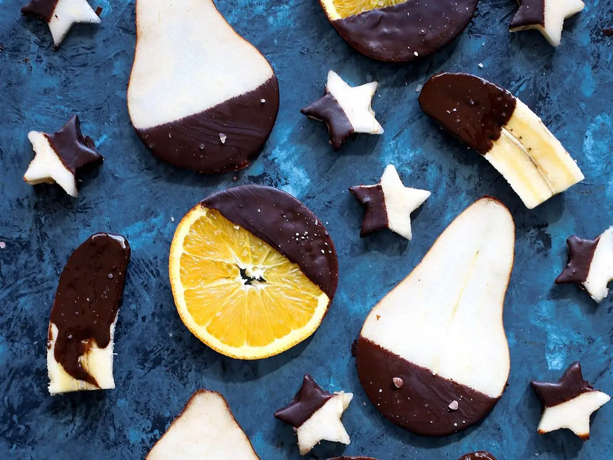 Gajos de pera, naranja y plátano bañados en chocolate negro sobre un fondo azul con estrellas
