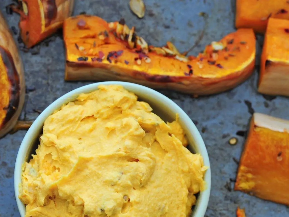 Cuenco de hummus anaranjado de calabaza rodeado de trozos de calabaza asada sobre una bandeja azul