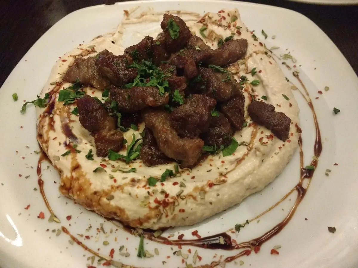 Plato de hummus cubierto de trozos de cordero especiado y perejil, con un hilo de salsa oscura
